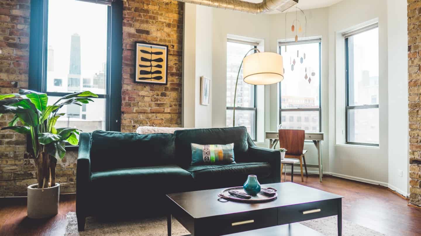 Photo of an apartment living room with a couch, coffee table, plant, lamp, and a work desk in the corner.