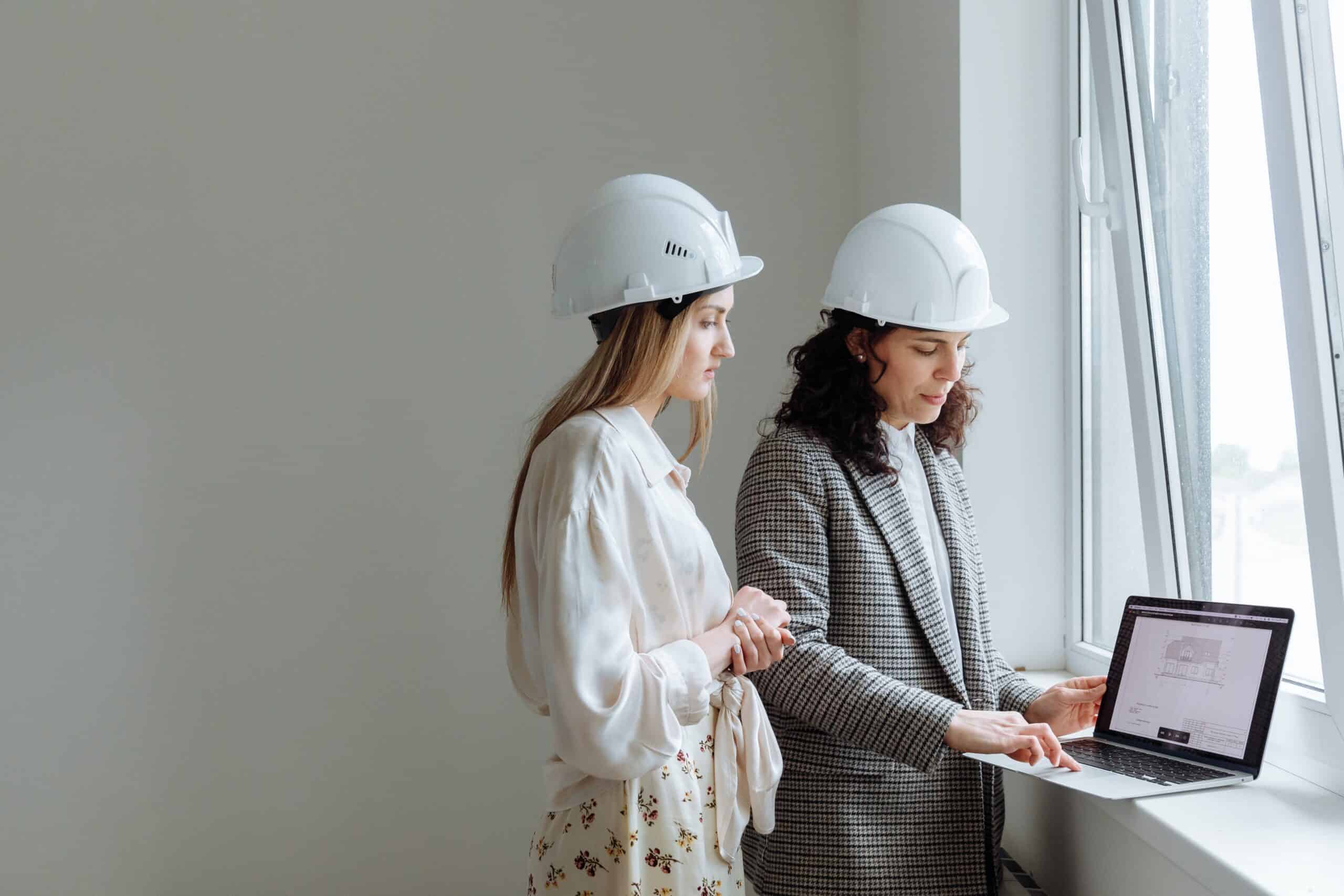 A photo of two women professionally dressed wearing hard hats and standing next to a window looking at a laptop screen.