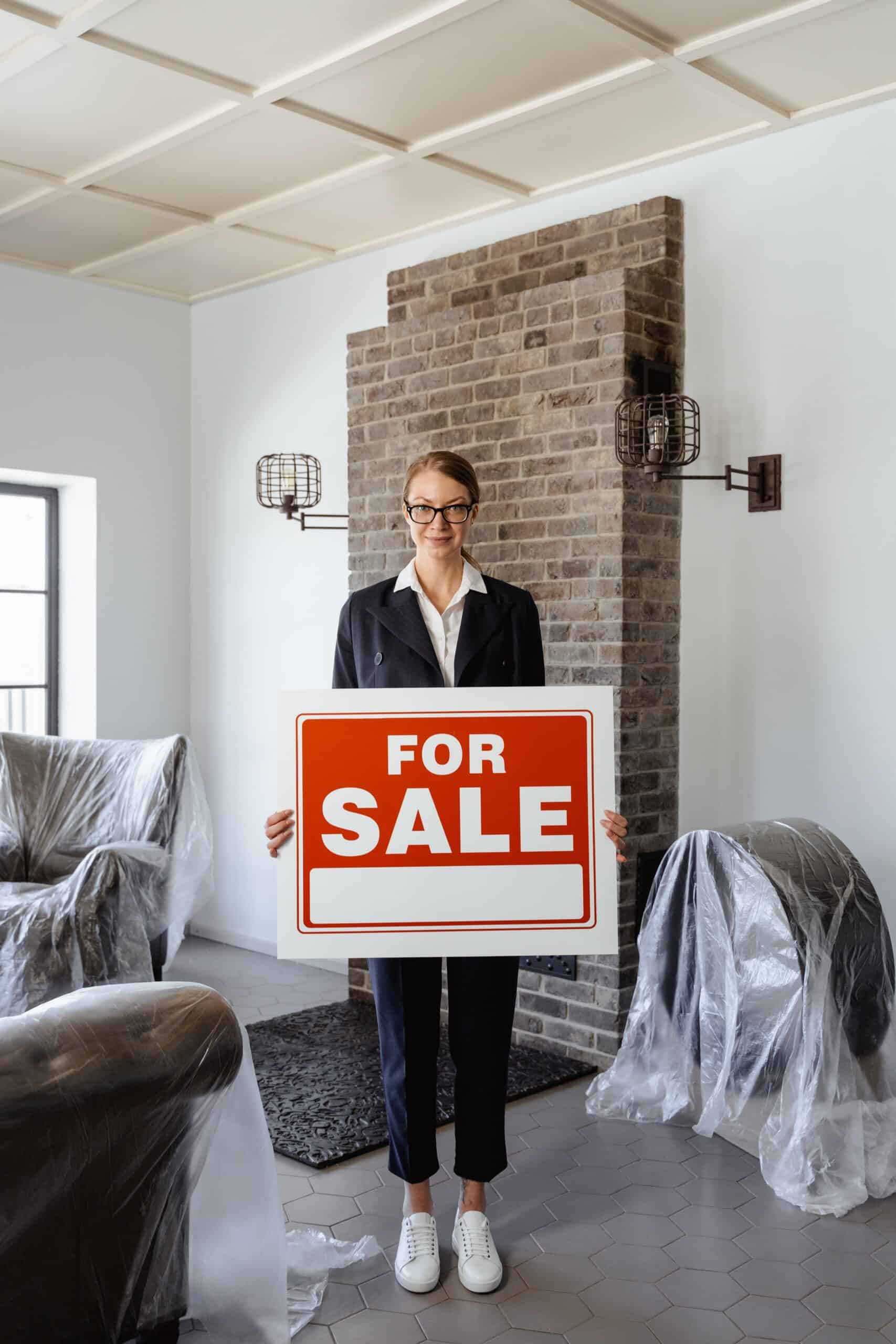 Woman smiling wearing glasses and a white shirt and blazer jacket holding a For Sale sign.