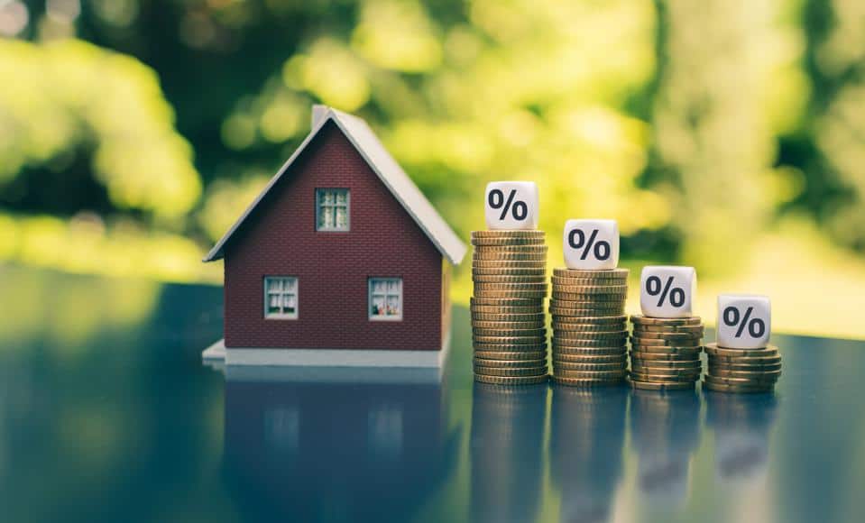 Photo of a house next to stacks of quarters and dice on top of the stacks. This is illustrating the cost of a home with interest rates and mortgages.